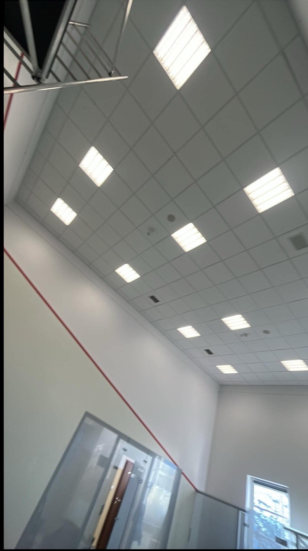 Wnętrze po remoncie: białe ściany z czerwoną linią, sufit z kwadratowymi lampami, szklana balustrada i okno. Widok z dołu, ukośny kadr.