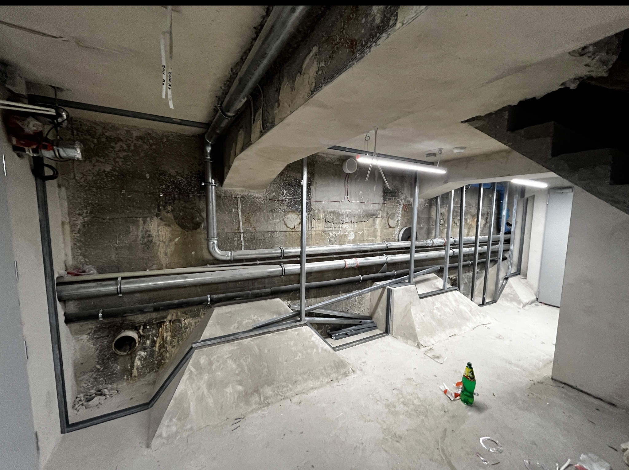 Instalacja rur i konstrukcji metalowych w surowym wnętrzu remontowanego mieszkania. Widoczne elementy instalacji pod sufitem i na ścianach, oraz fragment schodów.