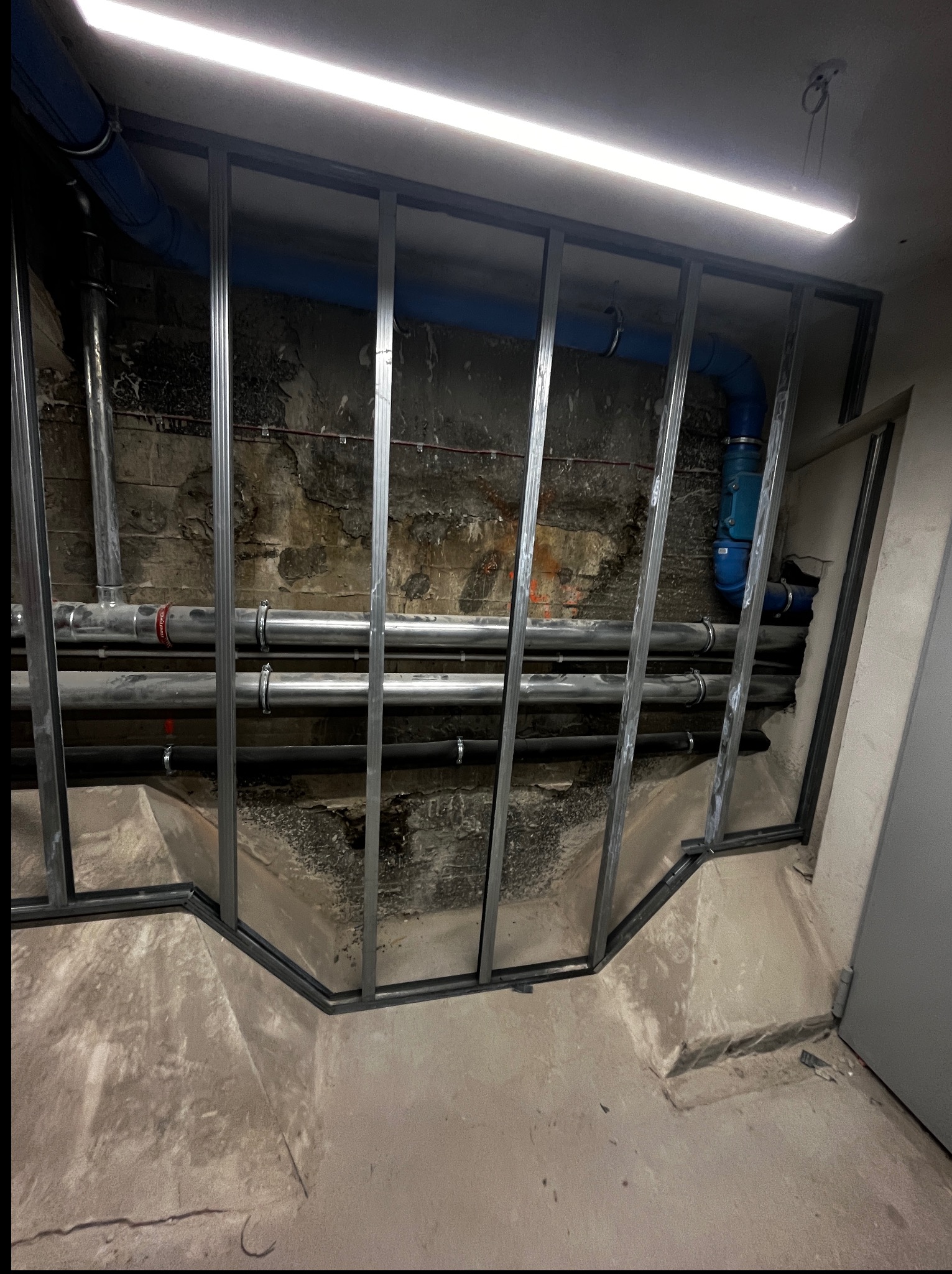 Metalowa konstrukcja szkieletowa na tle instalacji rur w piwnicy, widok z dołu. Etap przygotowawczy do zabudowy.