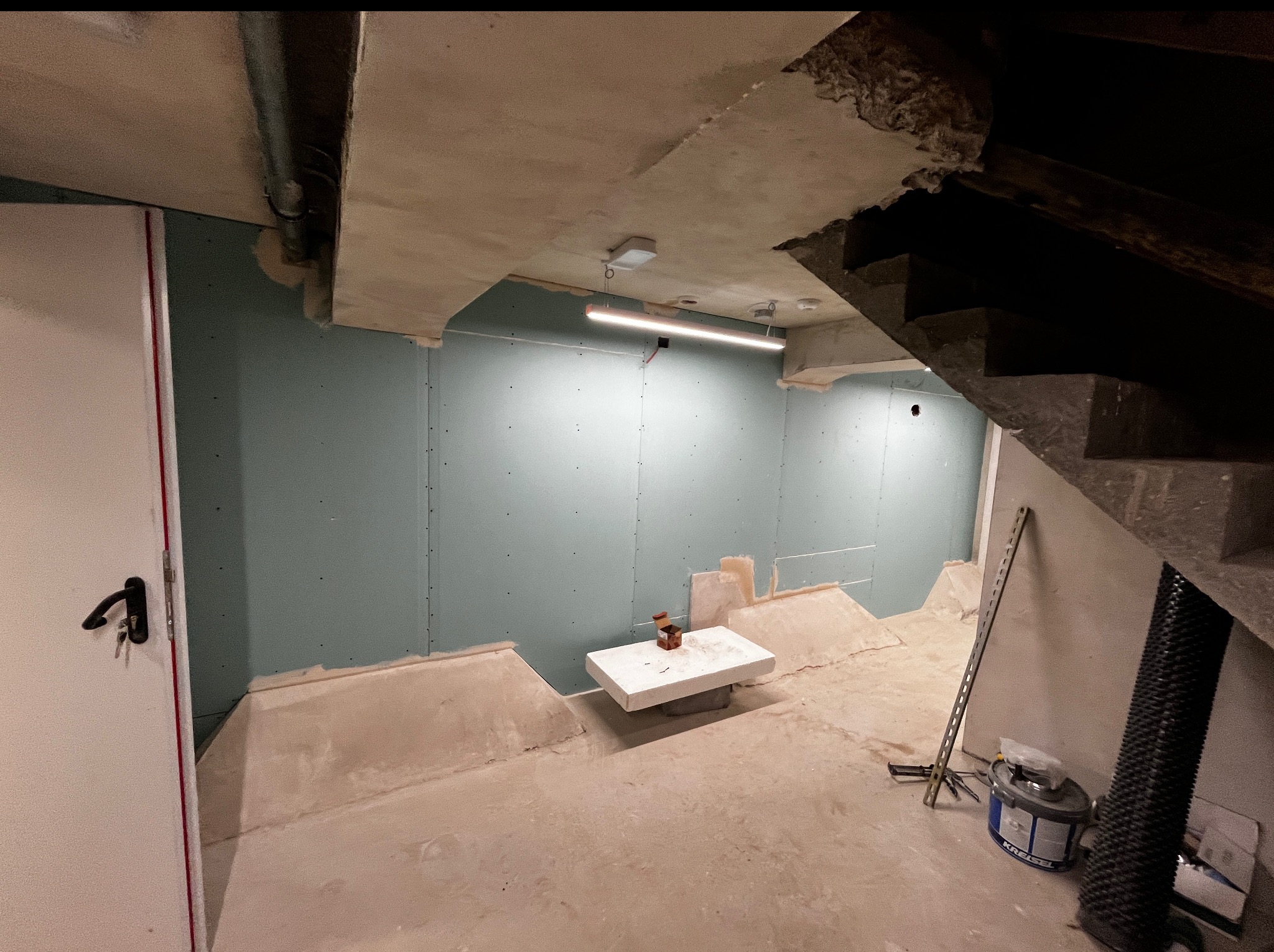 Remont piwnicy w trakcie: ściany z płyt gipsowych, surowy sufit z instalacjami, betonowa podłoga, schody i narzędzia. Widok na wnętrze w trakcie prac.