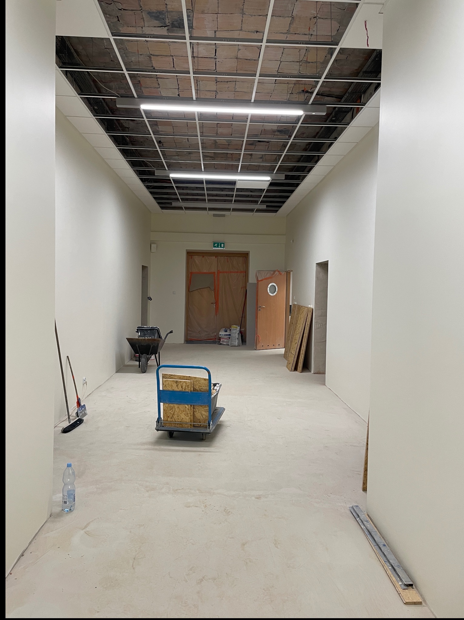 Wnętrze w trakcie remontu: surowy strop z instalacją, białe ściany, wózek z materiałami, taczka i butelka wody. Widok w perspektywie, podkreślający przestrzeń.
