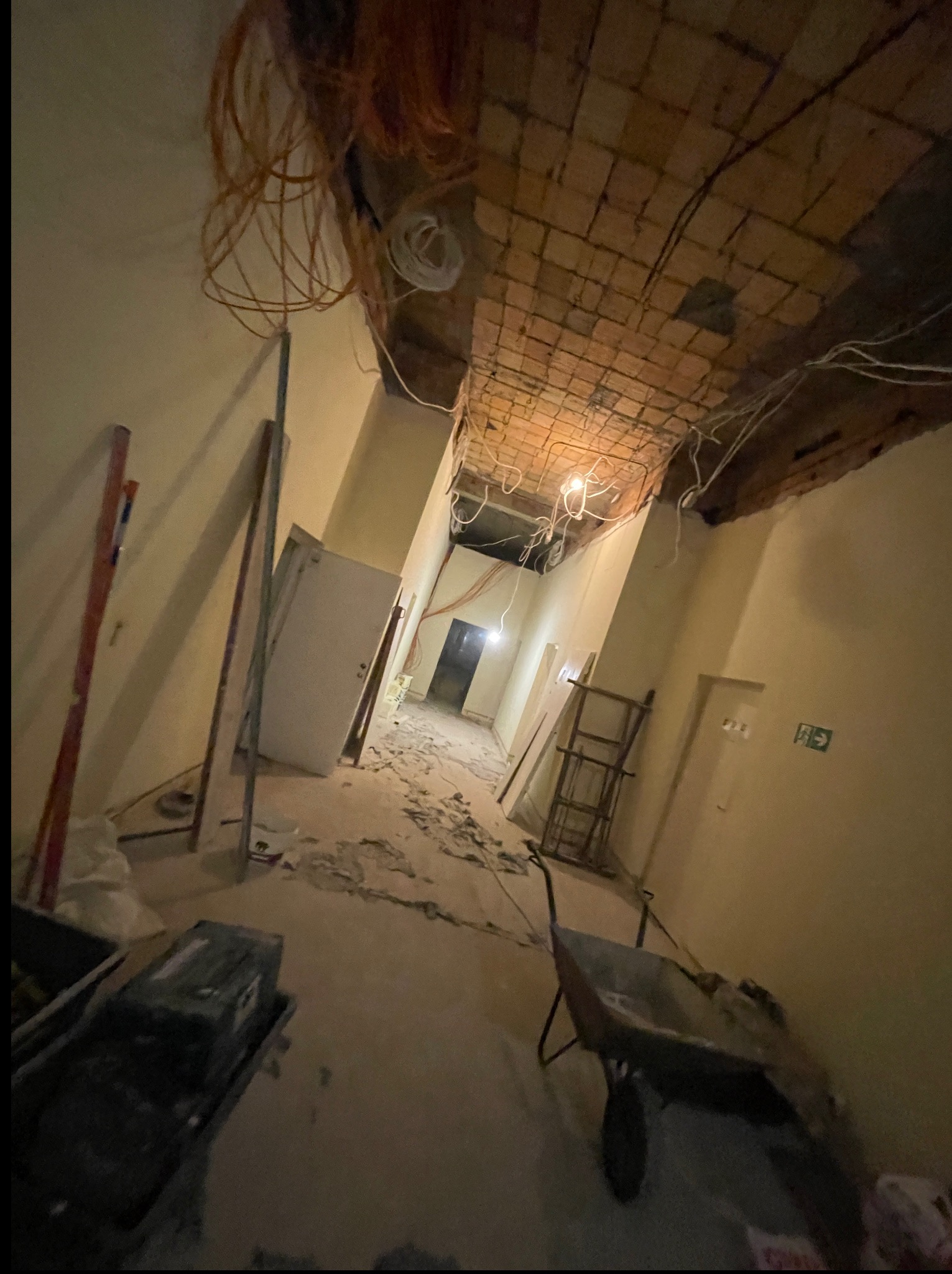 Wnętrze w trakcie remontu: odsłonięty ceglany strop, wiszące kable, narzędzia budowlane, gruz na podłodze. Widok z dołu, perspektywa zniekształcona.