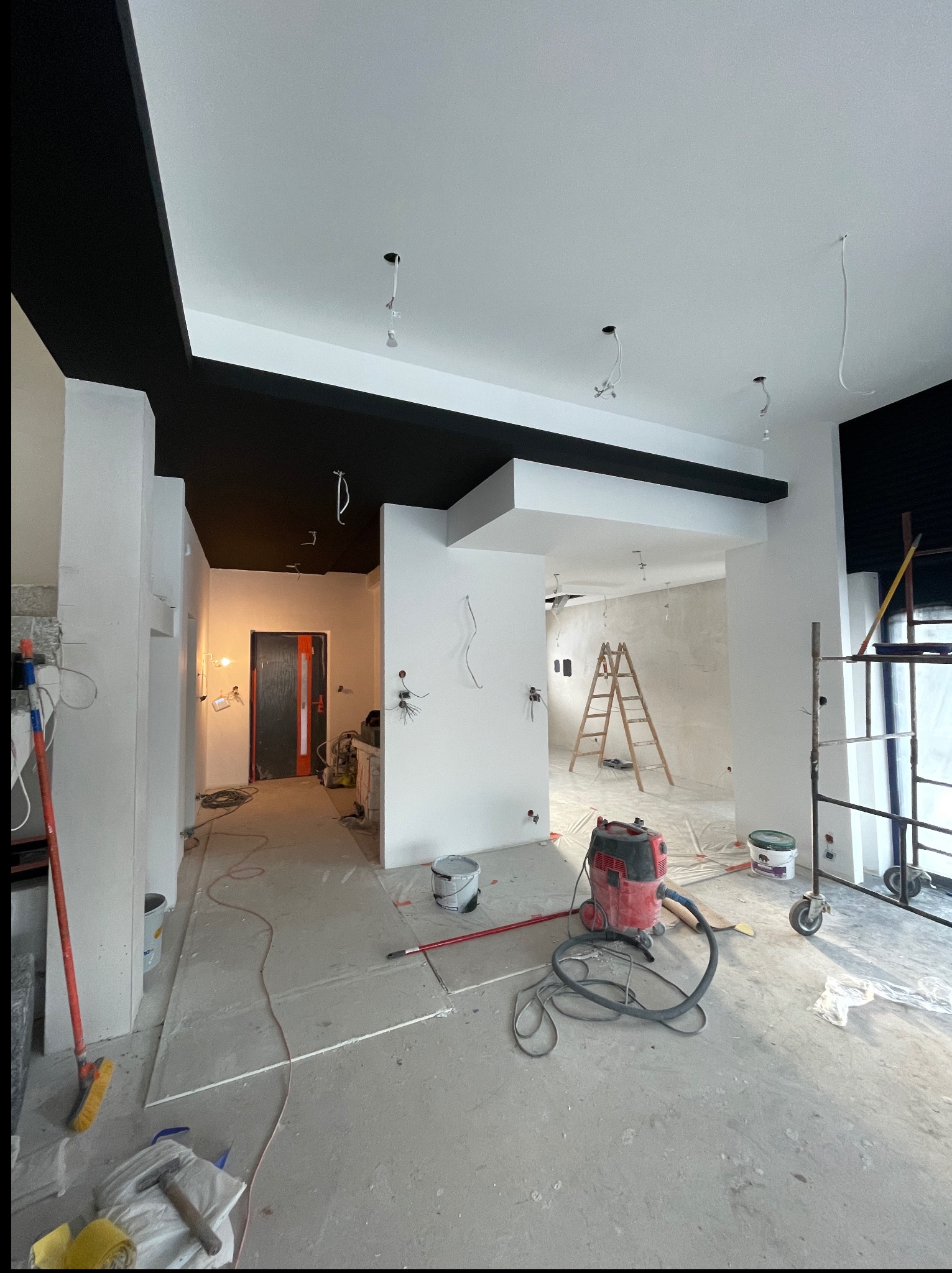 Wnętrze mieszkania w trakcie prac wykończeniowych: malowanie sufitu na czarno, białe ściany, drabina, sprzęt budowlany. Widoczne kable i puszki elektryczne.