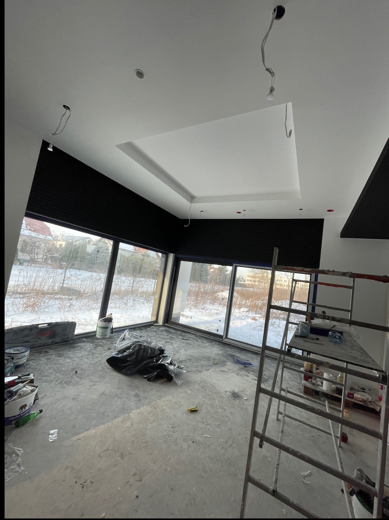 Wnętrze w trakcie wykończenia: surowe ściany, okna z roletami, rusztowanie. Widoczne kable oświetleniowe i materiały budowlane. Zimowy krajobraz za oknem.