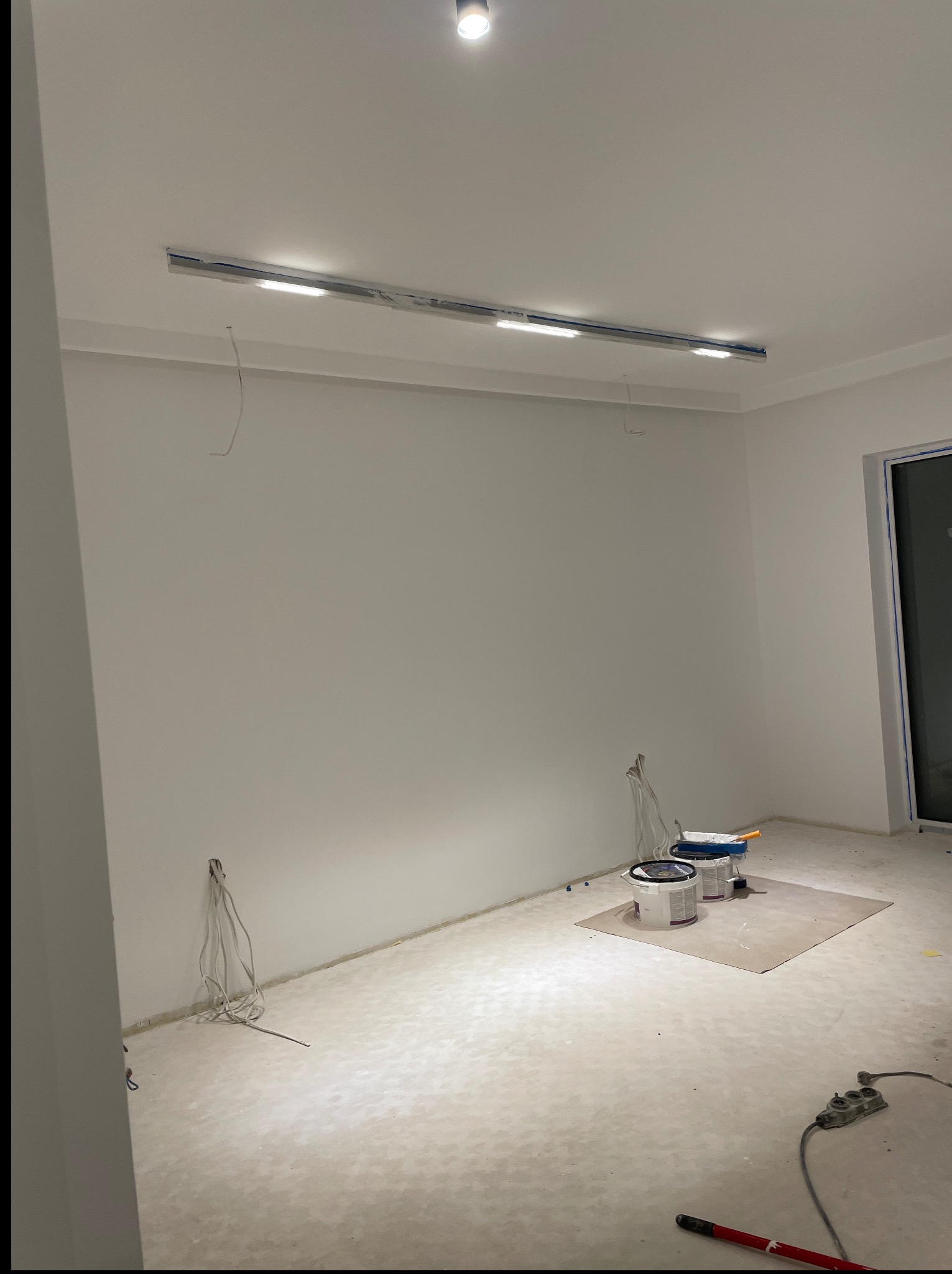 Pokój w trakcie malowania ścian na biało. Widoczne puszki z farbą, wałek malarski i listwa oświetleniowa pod sufitem. Kabel elektryczny wystaje ze ściany.