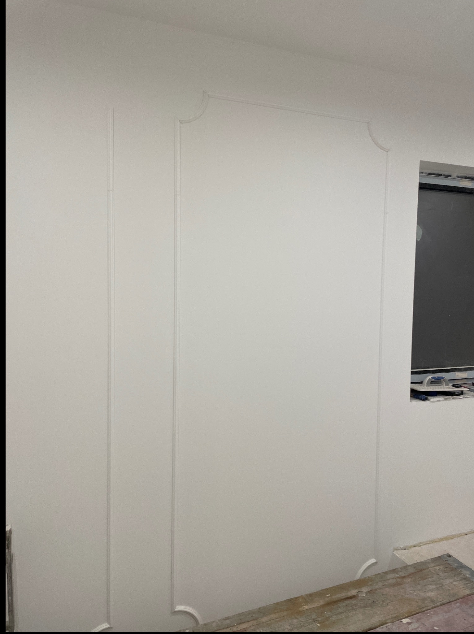 Biała ściana z dekoracyjnymi listwami przypodłogowymi i ramkami, świeżo po malowaniu. W tle widoczne okno i narzędzia malarskie.