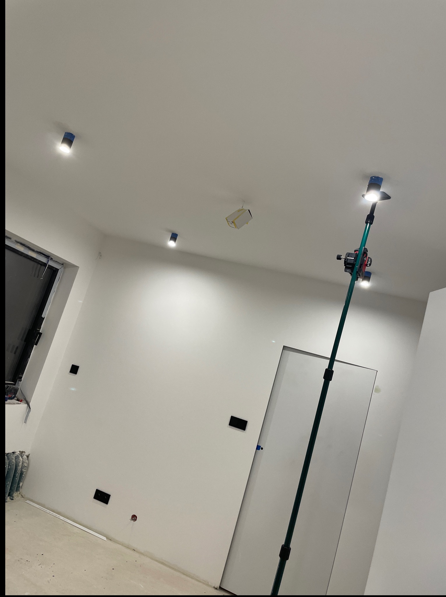 Białe wnętrze po remoncie z zamontowanymi lampami sufitowymi, widoczne gniazdka elektryczne i narzędzie do montażu oświetlenia na wysięgniku.