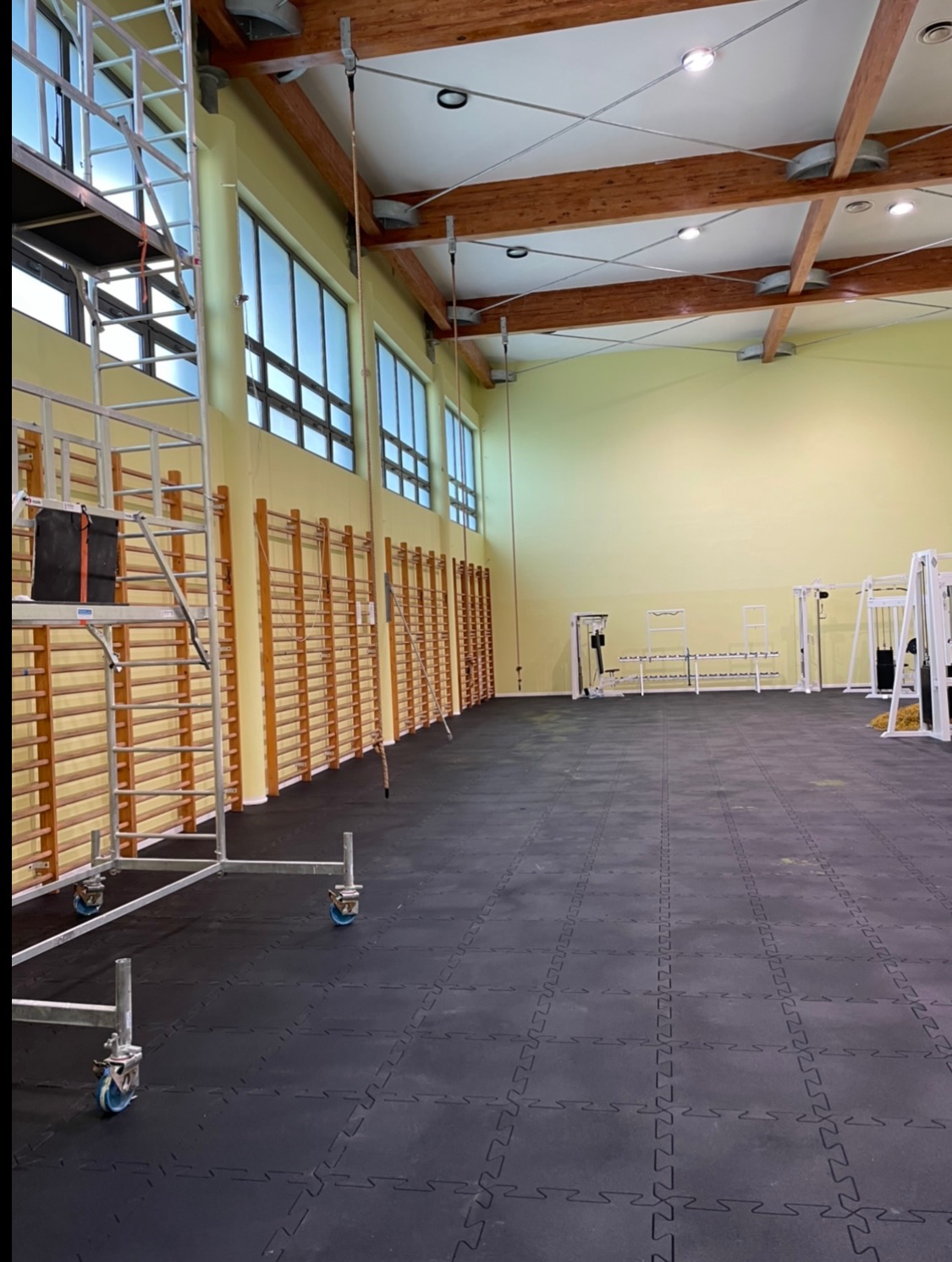 Sala gimnastyczna w trakcie remontu: rusztowanie na kółkach, drabinki gimnastyczne wzdłuż ściany, sprzęt do ćwiczeń w tle. Podłoga pokryta gumową nawierzchnią.