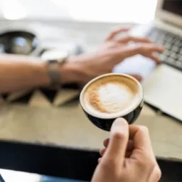 Osoba trzyma filiżankę cappuccino, w tle laptop i zegarek na nadgarstku, sugerujące przerwę w pracy lub spotkanie biznesowe w kawiarni.