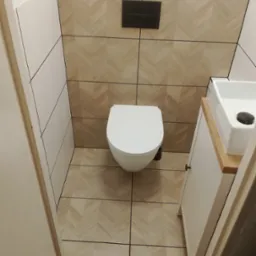 Nowoczesna toaleta w małej łazience z płytkami imitującymi drewno ułożonymi w jodełkę na ścianach i podłodze, podwieszana miska WC i umywalka nablatowa na szafce.