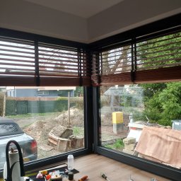 B&G SZKŁO KOSZALIN-SZKŁO NA WYMIAR GRZEGORZ GUŹNICZAK - Montaż drewnianych żaluzji w narożnym oknie z widokiem na ogród w trakcie prac porządkowych, narzędzia i akcesoria montażowe na blacie.