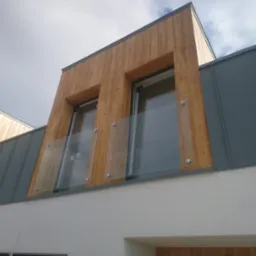 Nowoczesna elewacja budynku z drewnianą okładziną, szarą blachą i szklaną balustradą balkonową zamocowaną na metalowych wspornikach.