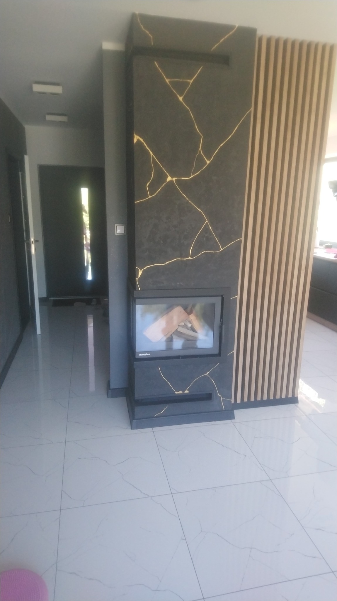 Nowoczesny salon z kominkiem obudowanym czarnym kamieniem z żyłkowaniem imitującym złoto, sąsiadującym z pionowymi panelami dekoracyjnymi z jasnego drewna.