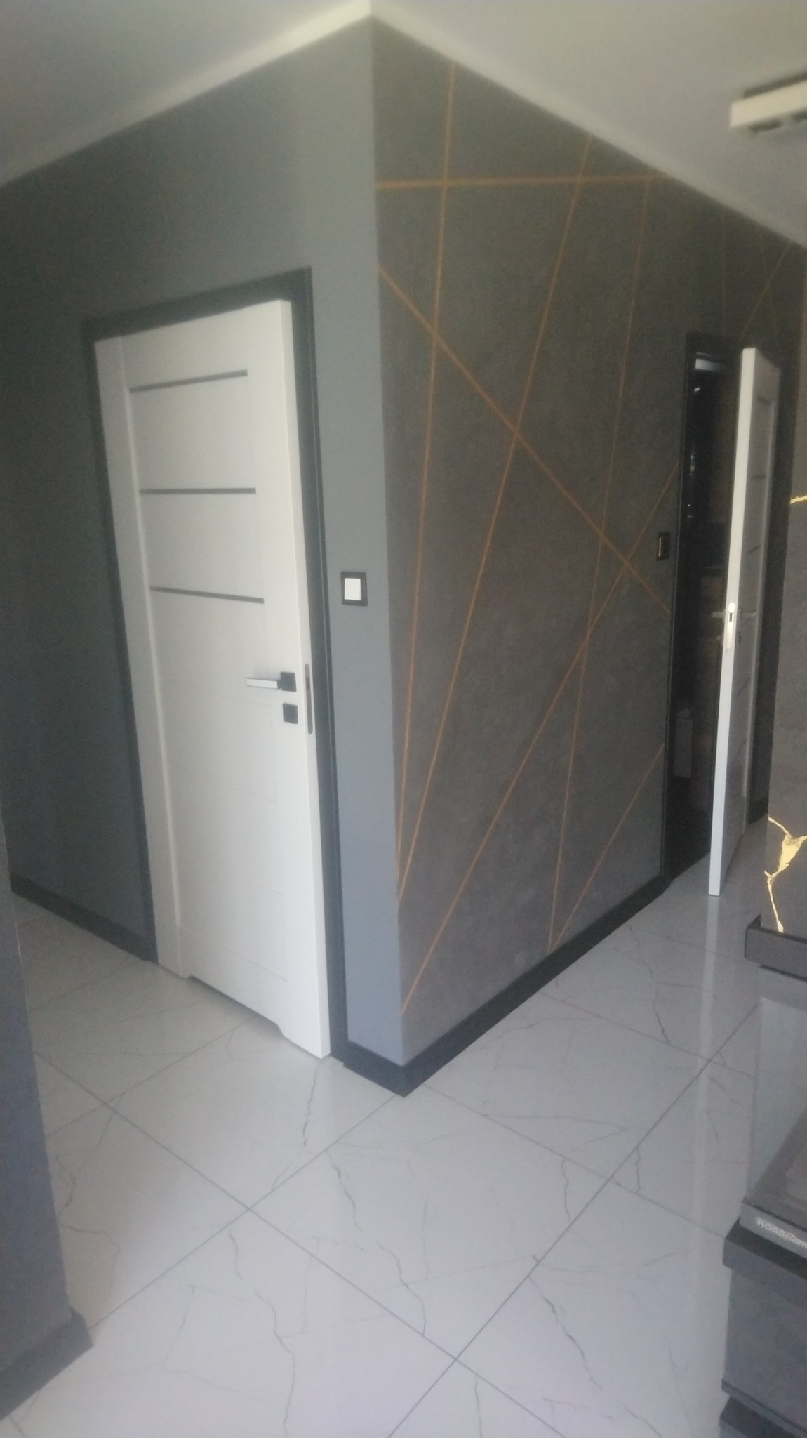 Nowoczesne wnętrze: szara ściana z geometrycznym wzorem w odcieniach złota, białe drzwi z czarną ościeżnicą i biała podłoga z marmurowym wzorem, widok z perspektywy narożnika.