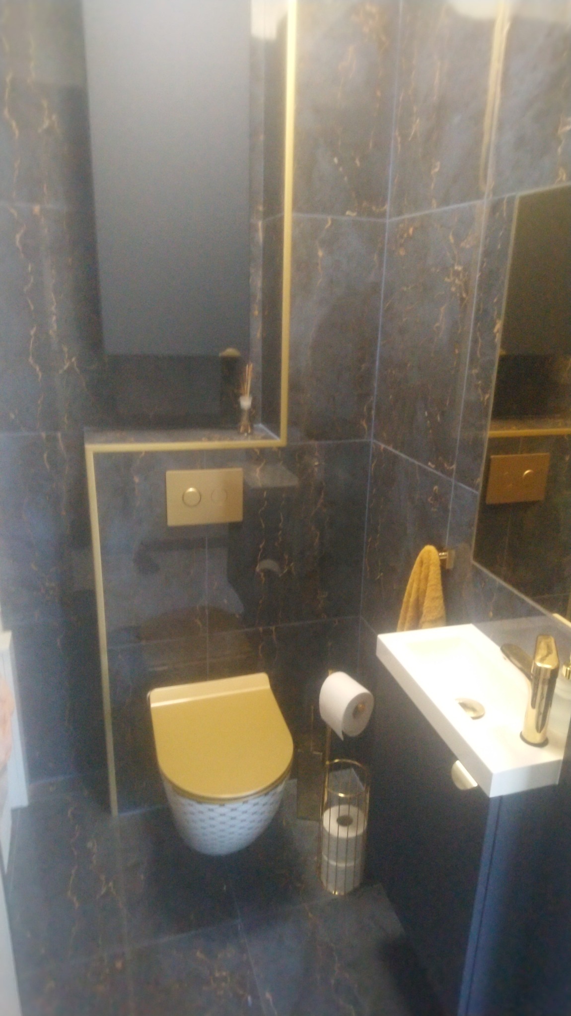 Ciemna łazienka z marmurowymi płytkami, złotymi akcentami i podwieszaną toaletą z deską w kolorze miodowym oraz białą umywalką.