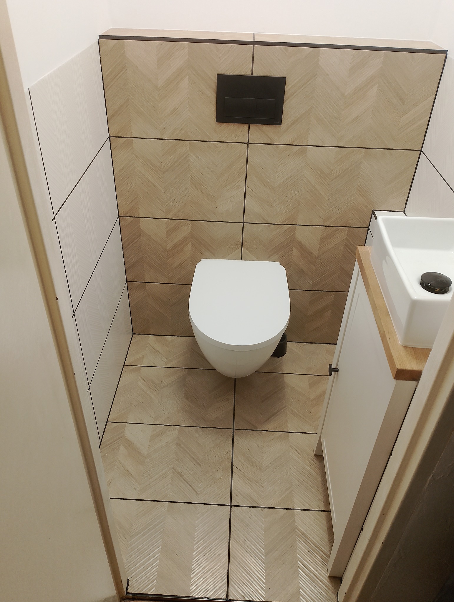 Nowoczesna toaleta w małej łazience z płytkami imitującymi drewno ułożonymi w jodełkę na ścianach i podłodze, podwieszana miska WC i umywalka nablatowa na szafce.