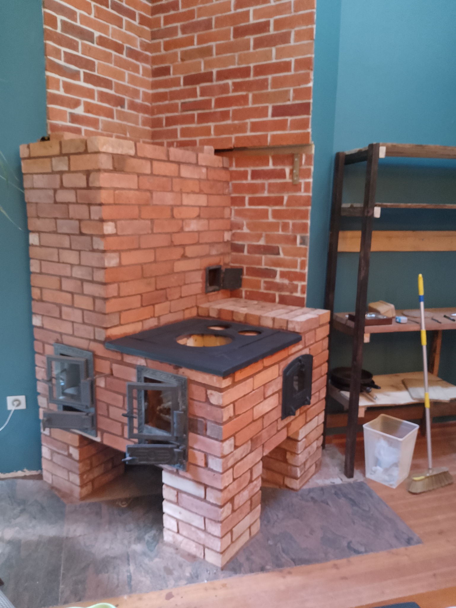 Murowany piec kuchenny z cegły, z otwartymi żeliwnymi drzwiczkami paleniska i czarnym blatem z otworami na garnki, na tle niebieskiej ściany i drewnianego regału.