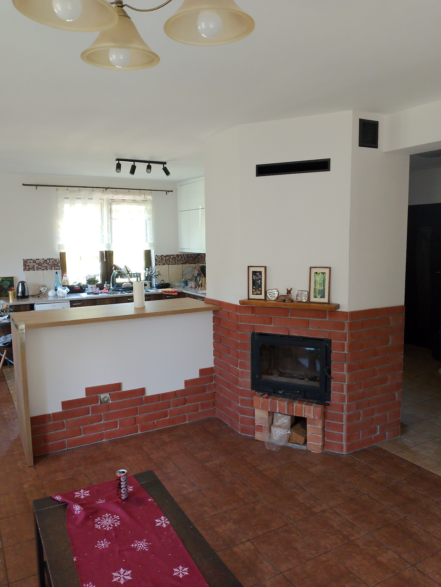 Częściowy widok salonu z ceglanym kominkiem, drewnianym blatem oddzielającym kuchnię, brązowymi płytkami na podłodze i stolikiem kawowym z czerwoną serwetą w śnieżynki.