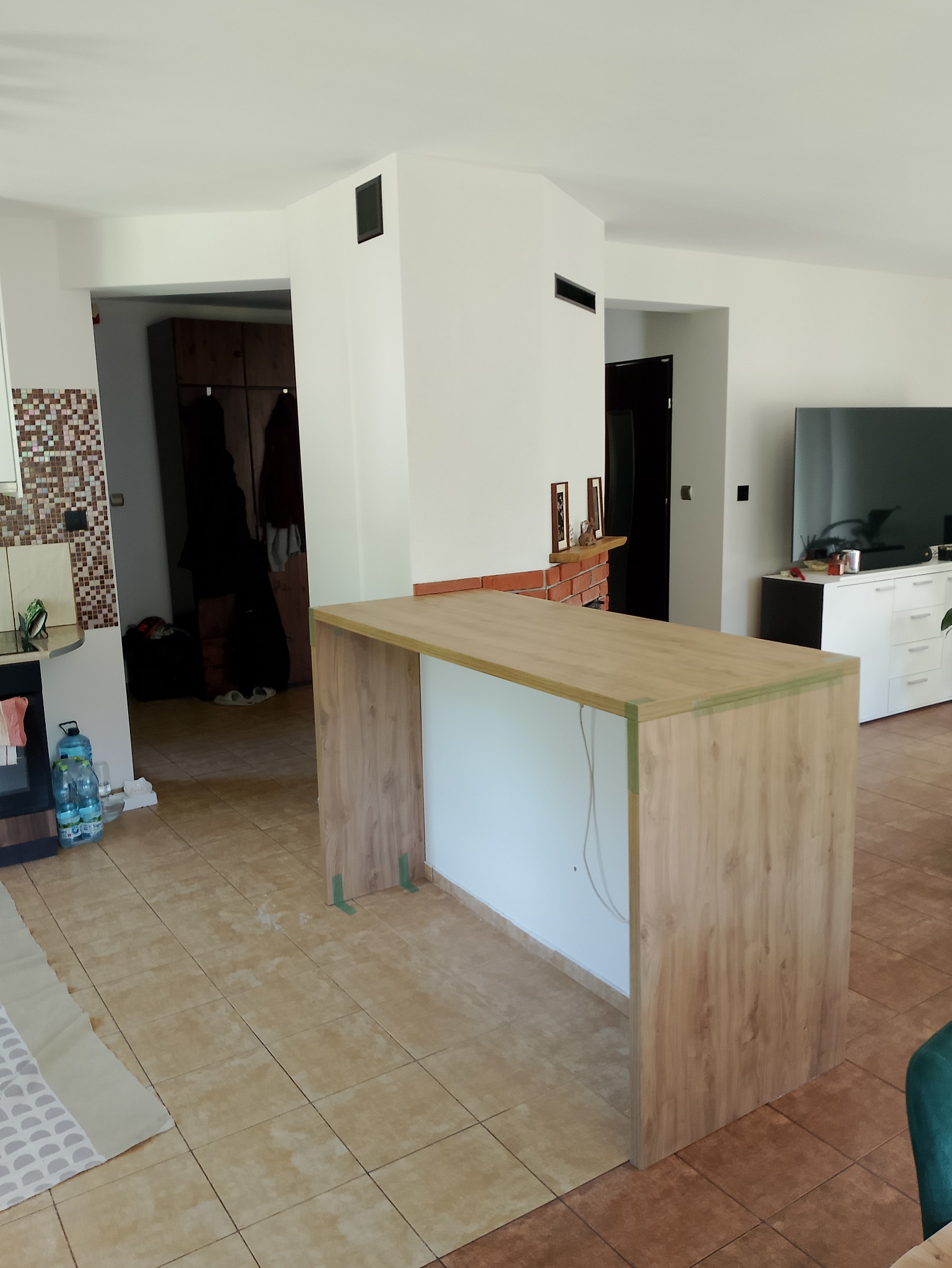 Wnętrze w trakcie prac wykończeniowych: surowa wyspa kuchenna z drewnopodobnym blatem i okleiną, zabezpieczona zieloną taśmą, na tle częściowo wykończonego pomieszczenia z terakotą na podłodze...