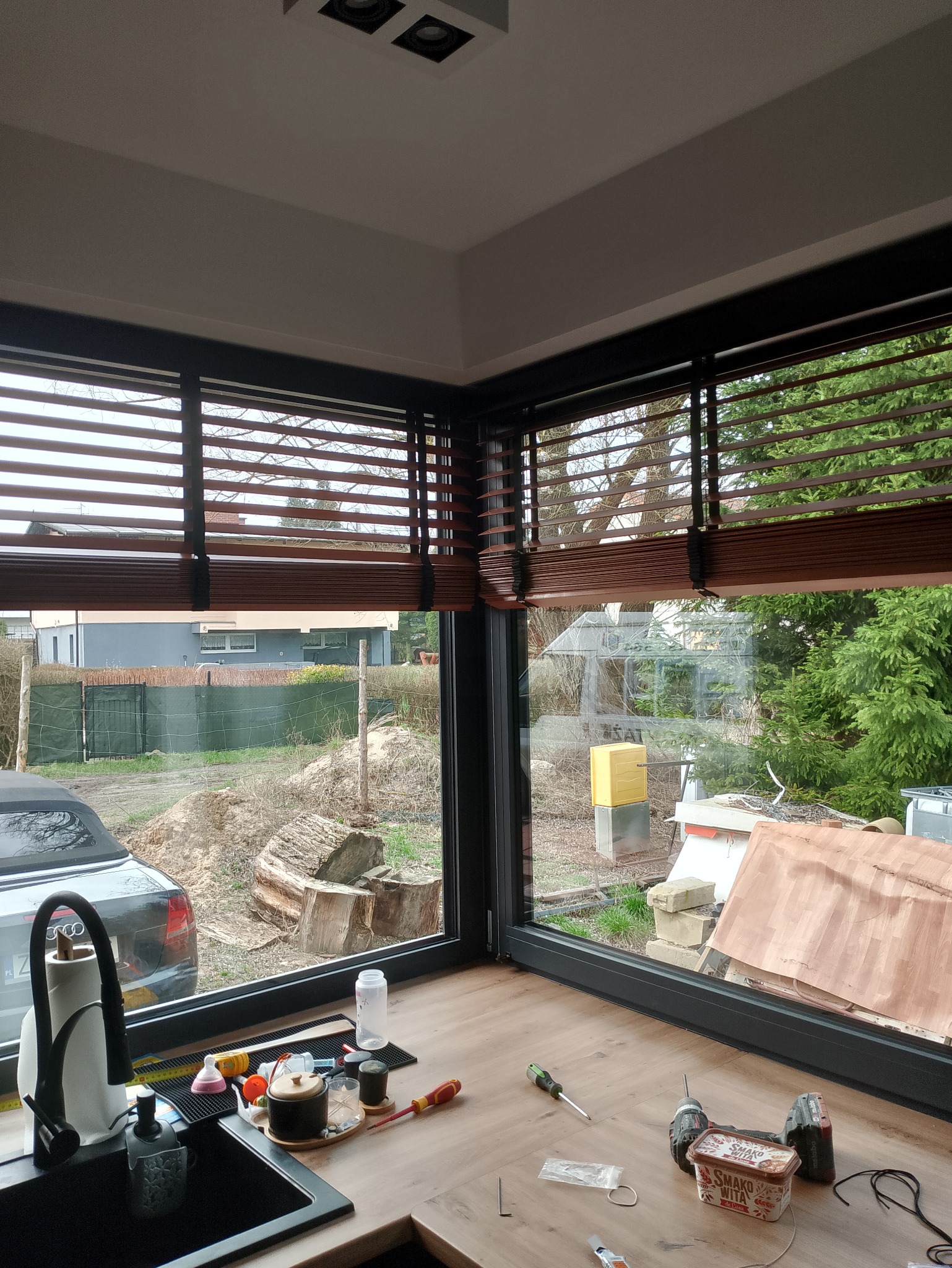 Montaż drewnianych żaluzji w narożnym oknie z widokiem na ogród w trakcie prac porządkowych, narzędzia i akcesoria montażowe na blacie.