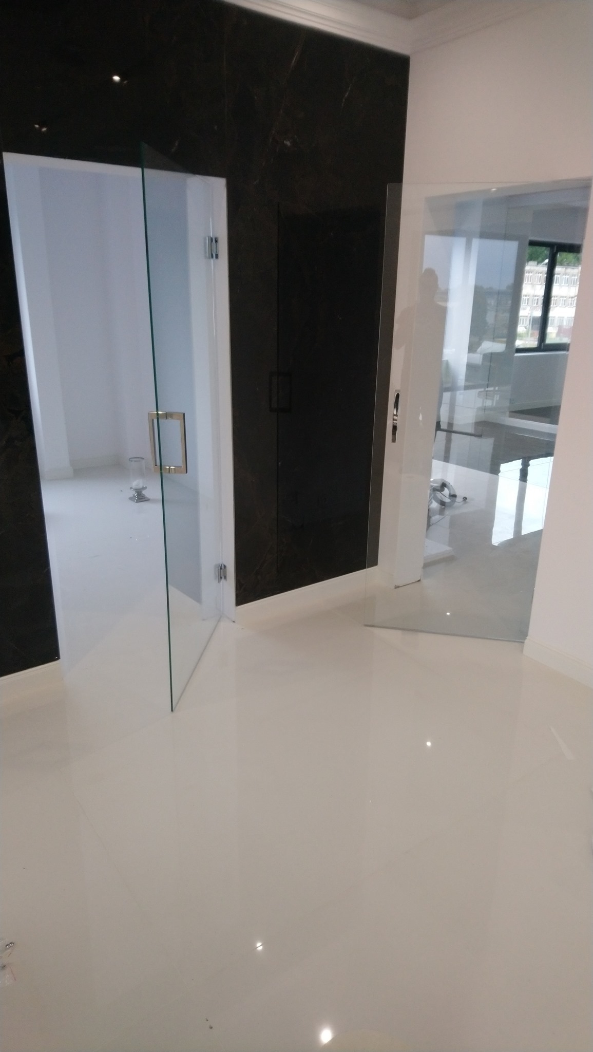 Nowoczesne, przeszklone drzwi w jasnym pomieszczeniu z błyszczącą, kremową podłogą i ciemną, marmurową ścianą. Widoczne metalowe okucia i uchwyt drzwiowy. W tle widok na budynek przez szybę.