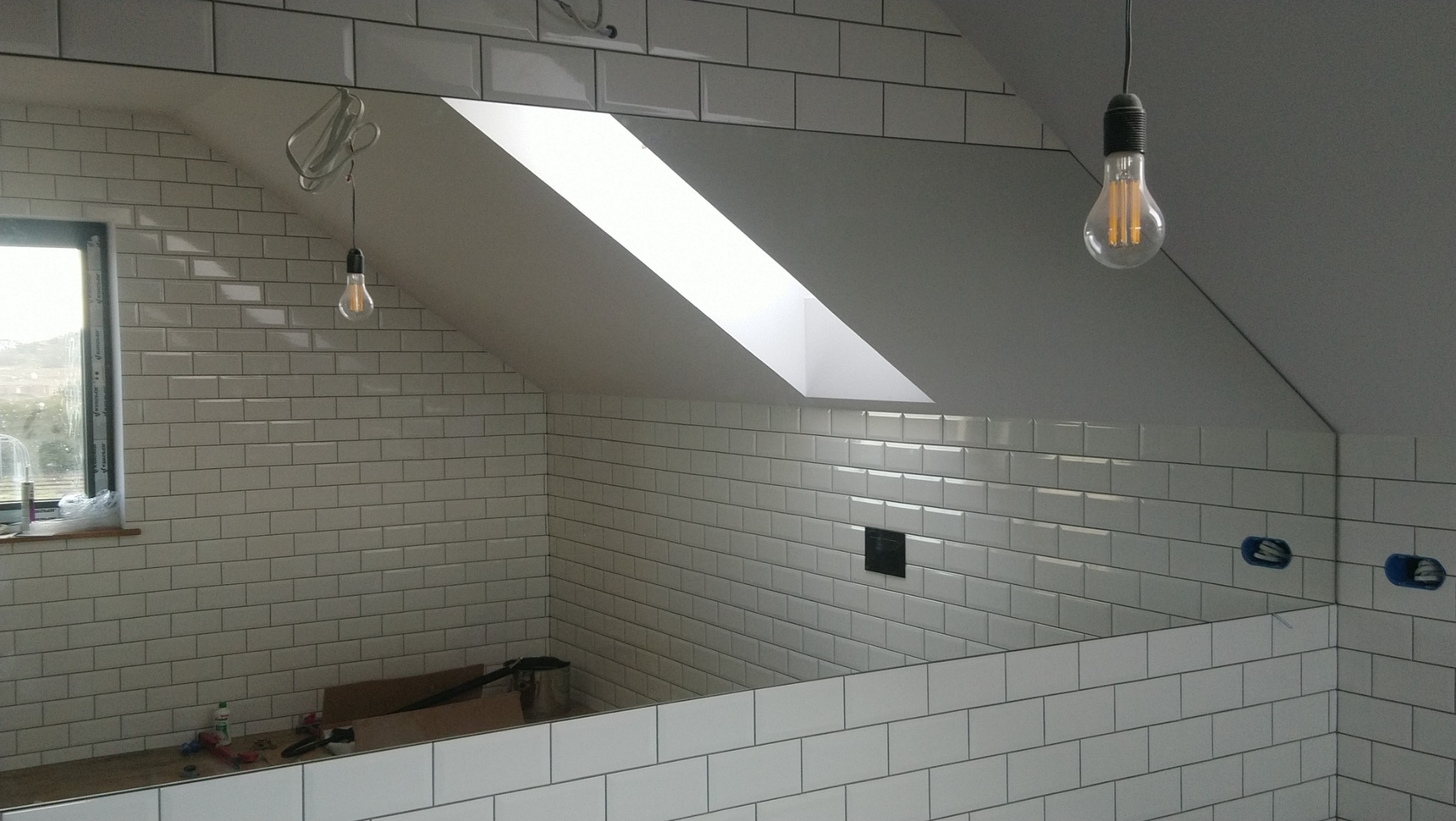 Ściany i skośny sufit w trakcie wykańczania białymi, prostokątnymi płytkami, widoczne okno dachowe, wiszące żarówki dekoracyjne i puszki elektryczne w ścianie.