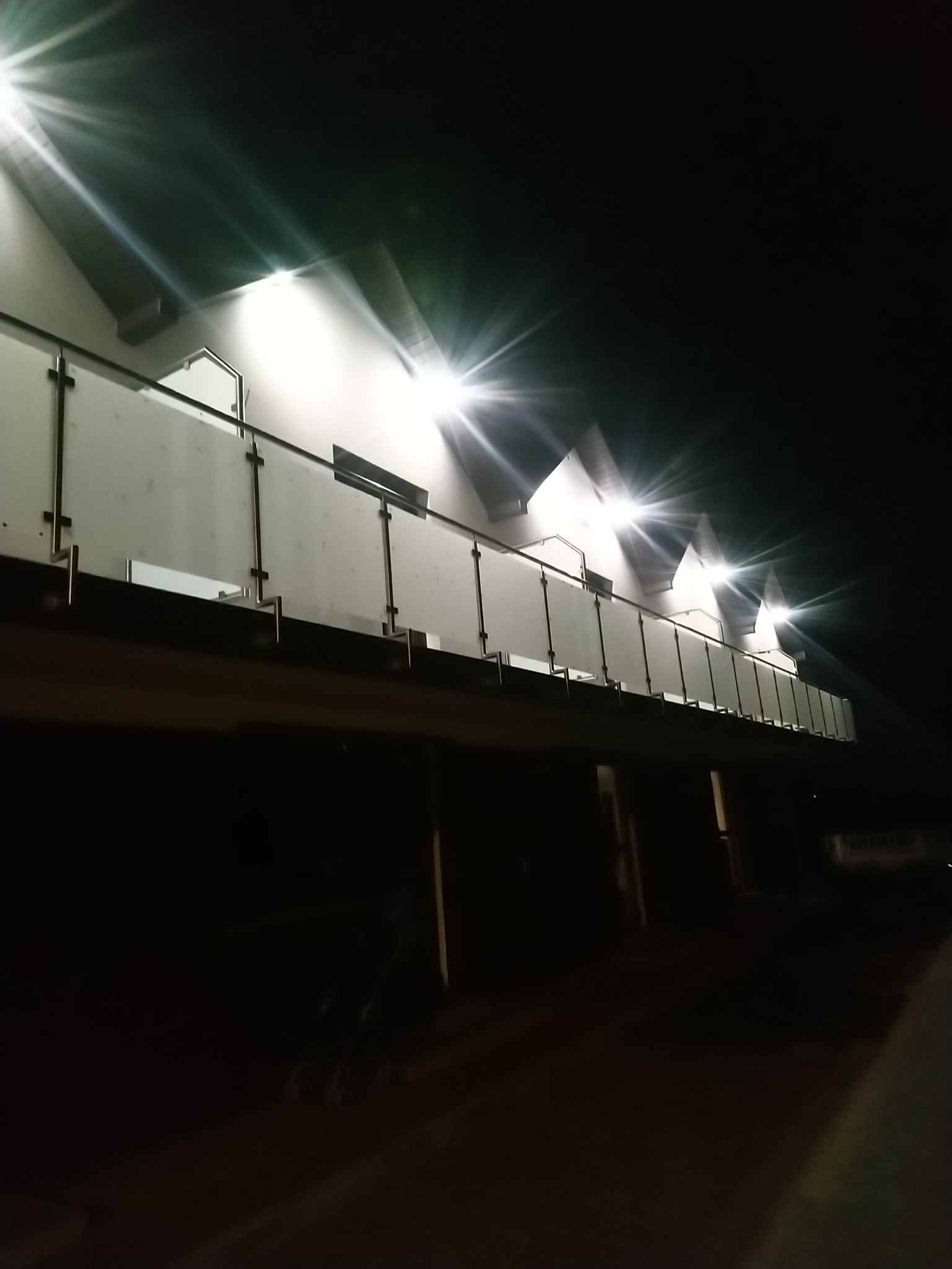 Nocny widok z dołu na balkon z mleczną balustradą szklaną i metalowymi poręczami, oświetlony punktowymi reflektorami, budynek w tle.