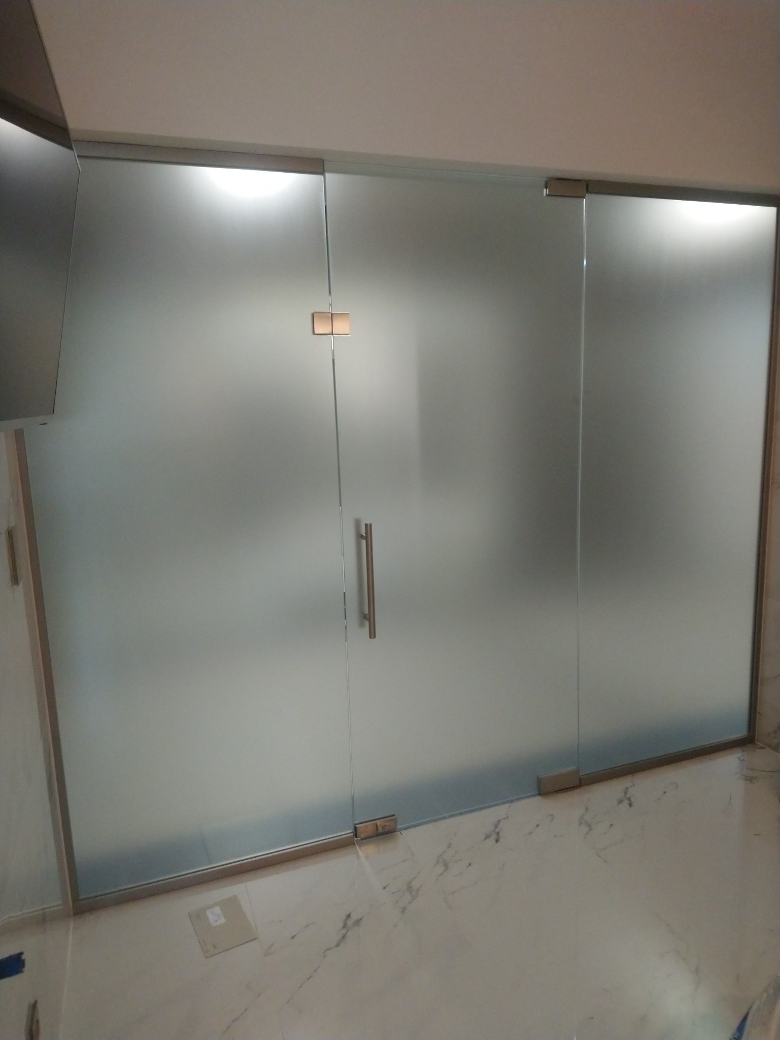 Szklane drzwi przesuwne z matowego szkła, wyposażone w minimalistyczny uchwyt i widoczne okucia, zamontowane w pomieszczeniu z marmurową podłogą.