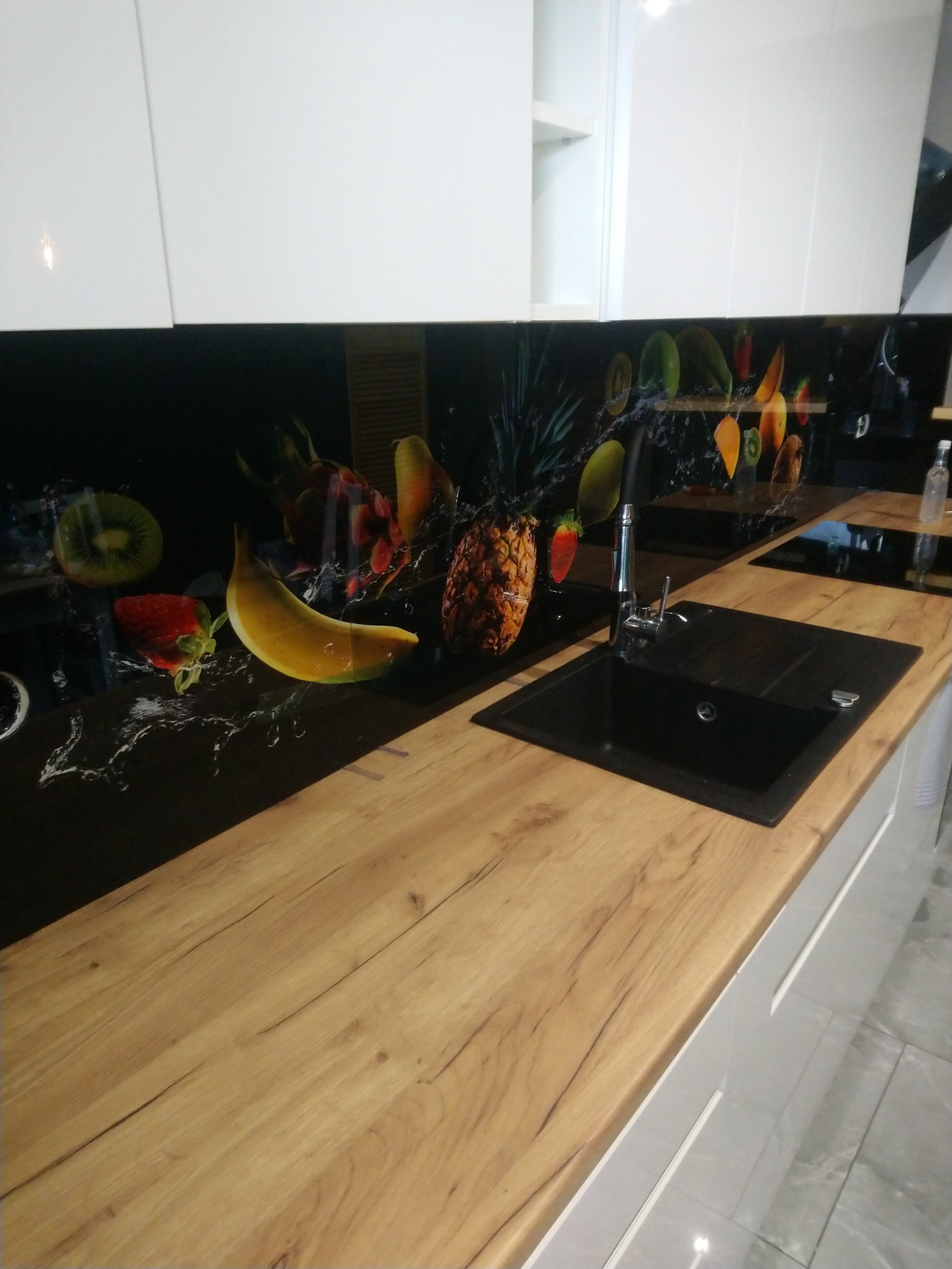 Kuchnia z drewnianym blatem, czarnym zlewem i szklanym panelem ściennym z motywem owoców.