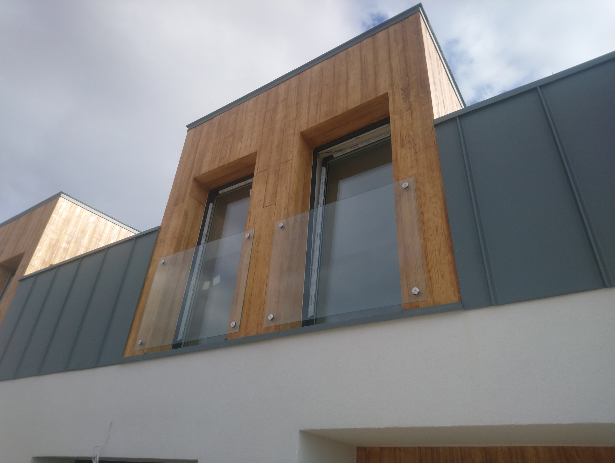 Nowoczesna elewacja budynku z drewnianą okładziną, szarą blachą i szklaną balustradą balkonową zamocowaną na metalowych wspornikach.