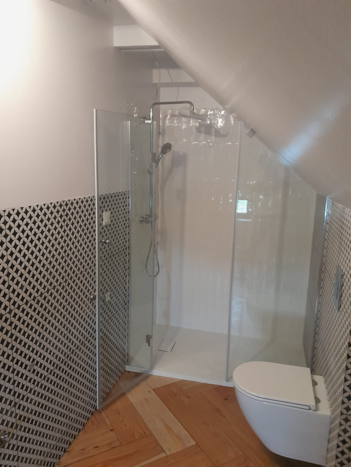 Nowoczesna kabina prysznicowa ze szklanymi drzwiami w łazience na poddaszu, białe płytki imitujące pionowe panele, wzorzyste płytki na ścianie, drewniana podłoga ułożona w jodełkę.