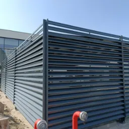 Nowoczesne, poziome, metalowe ogrodzenie lamelowe w kolorze grafitowym, z widocznymi elementami instalacji przeciwpożarowej w postaci czerwonych hydrantów i żwirowym podłożem.