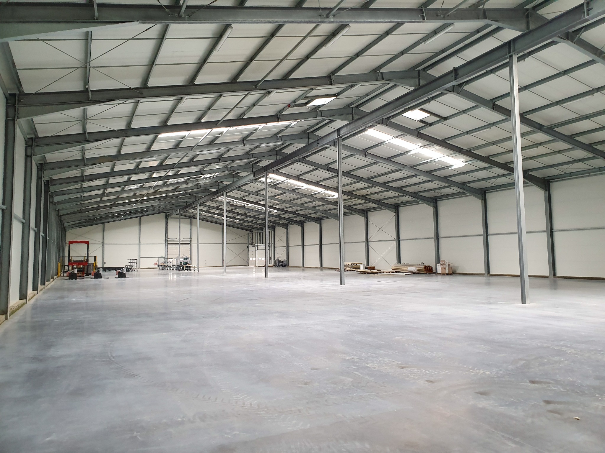 Wnętrze nowo wybudowanej, przestronnej hali z szarą, betonową posadzką, stalową konstrukcją nośną i białymi ścianami z paneli warstwowych, oświetlonej przez okna dachowe i lampy.