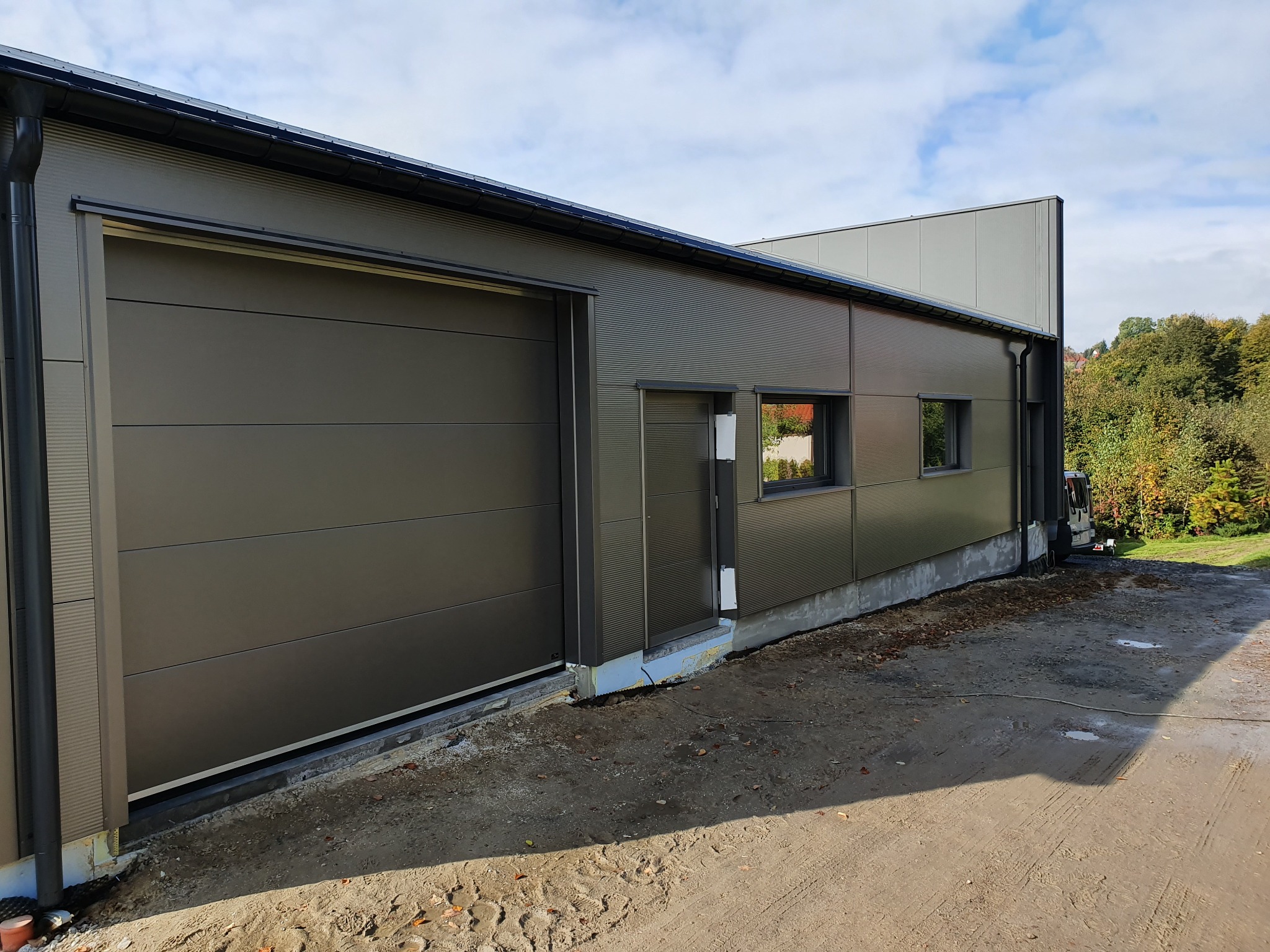 Nowoczesna hala z szarej blachy trapezowej, z częściowo otwartą bramą segmentową, widoczne okna i drzwi wejściowe, teren wokół w trakcie prac wykończeniowych.