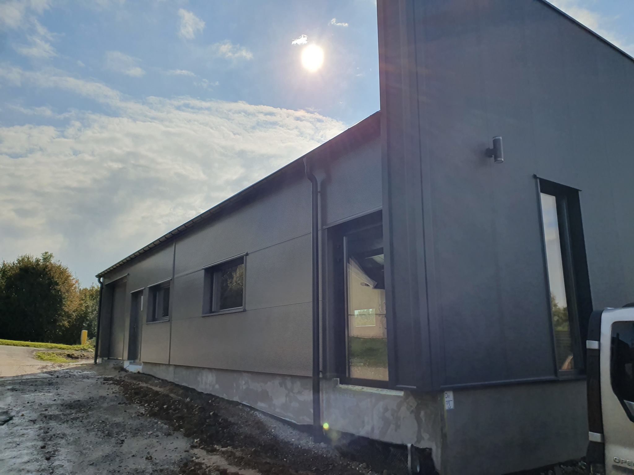 Nowoczesna hala przemysłowa w trakcie budowy, widok z zewnątrz, z widocznymi panelami warstwowymi i oknami, na tle błękitnego nieba z chmurami, świecące słońce.