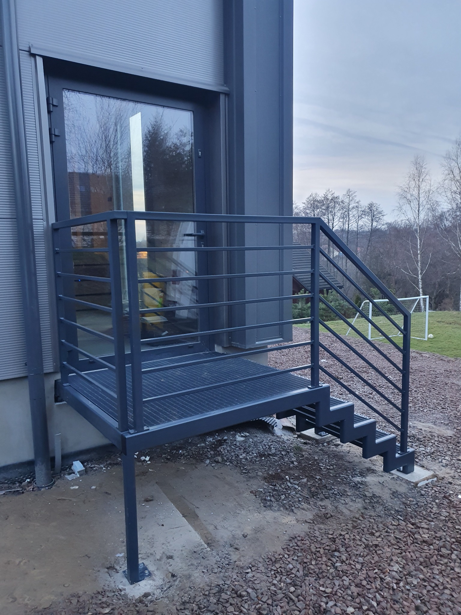 Zewnętrzne metalowe schody z podestem, prowadzące do drzwi budynku w Rzeszotarach, widoczne z boku, z piłką i bramką w tle.