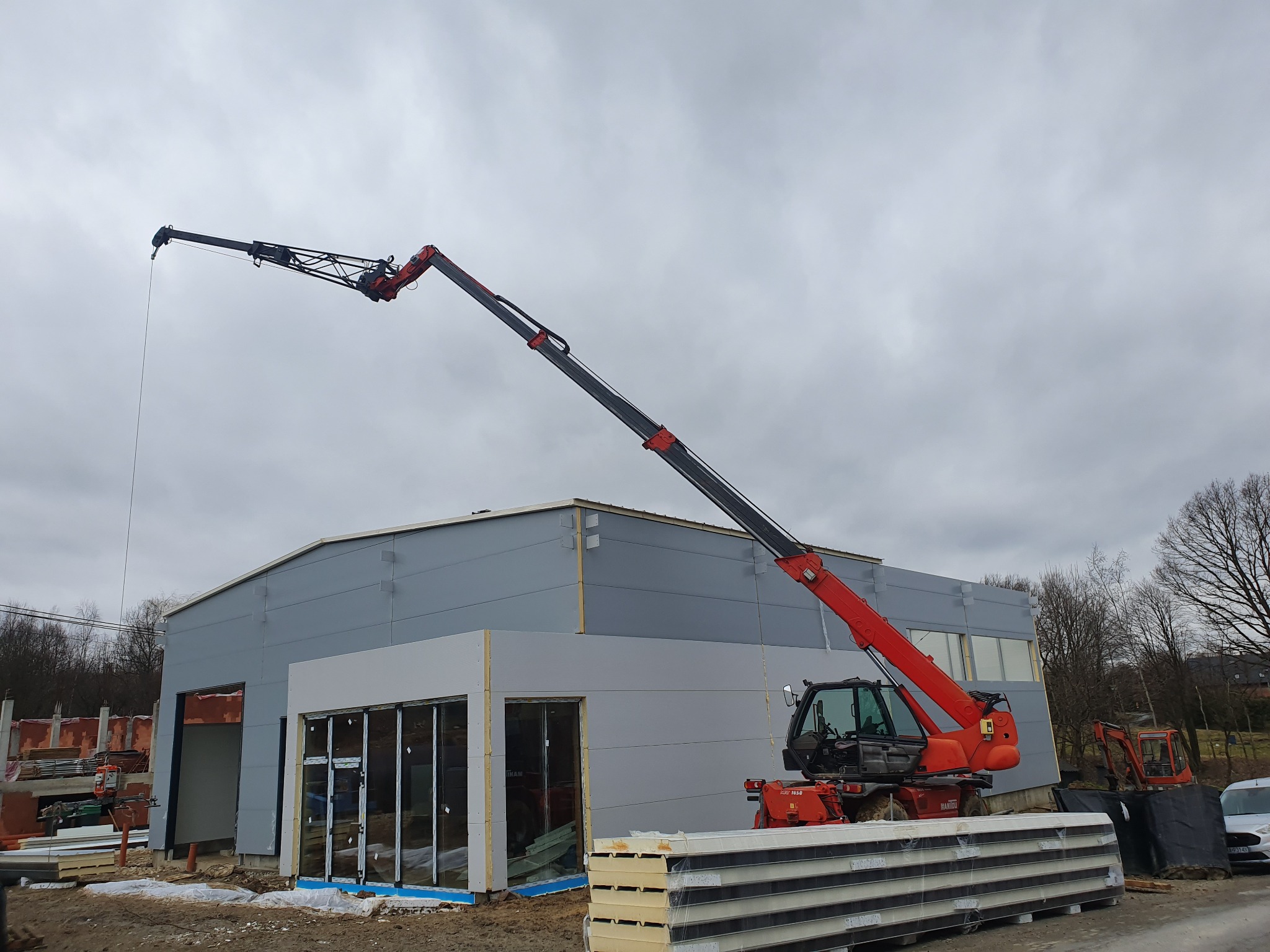 Czerwony podnośnik teleskopowy Manituo obsługuje montaż szarych płyt warstwowych na ścianie powstającej hali; w tle widoczne rusztowania i mini koparka.