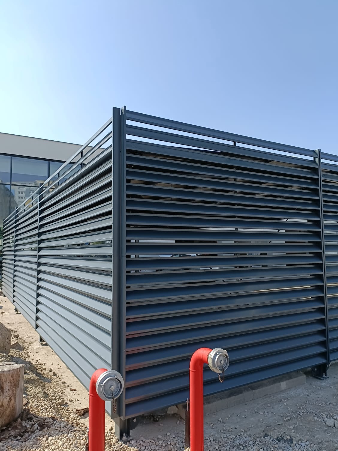Nowoczesne, poziome, metalowe ogrodzenie lamelowe w kolorze grafitowym, z widocznymi elementami instalacji przeciwpożarowej w postaci czerwonych hydrantów i żwirowym podłożem.