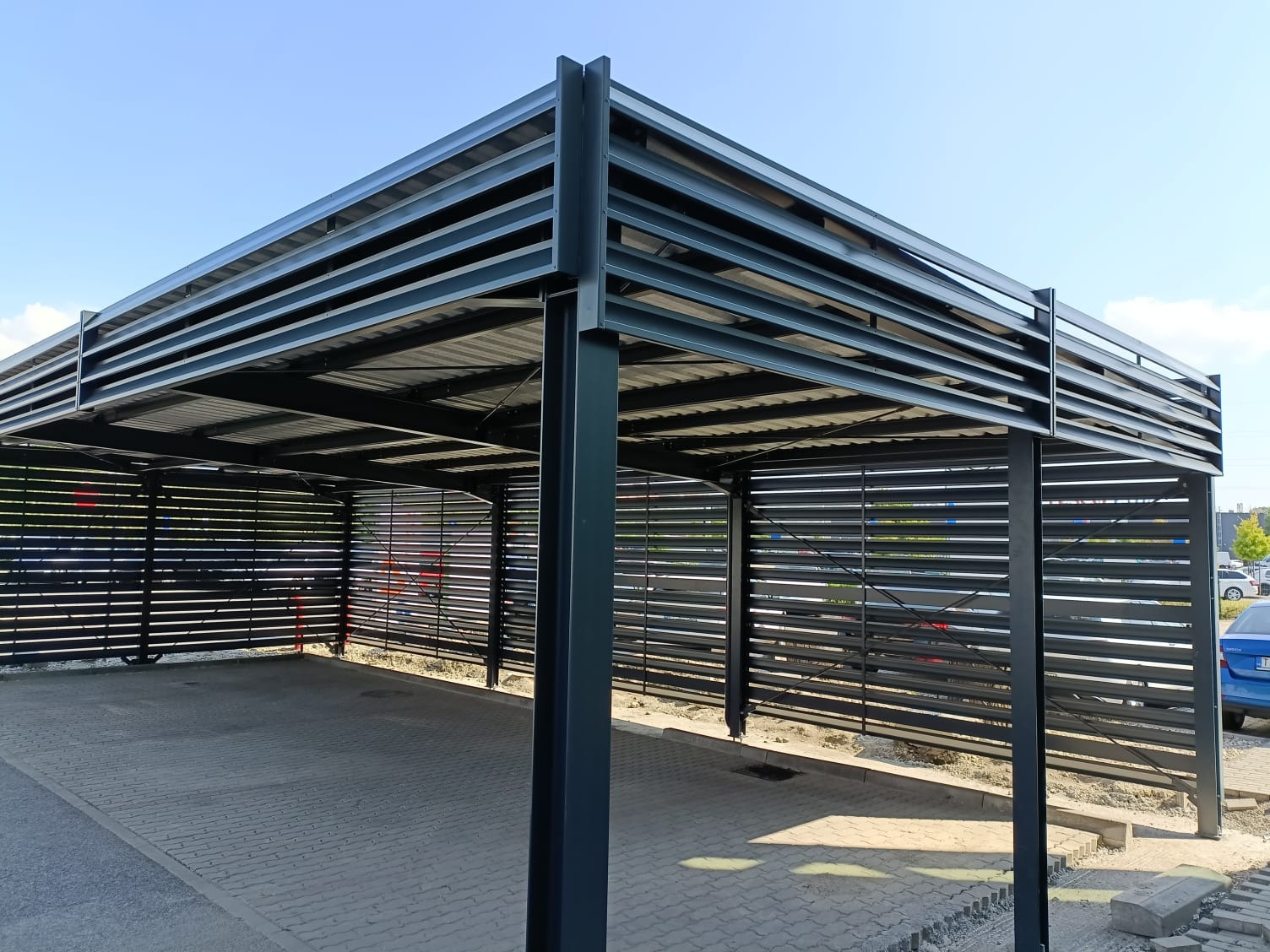 Nowoczesna, metalowa wiata parkingowa z poziomymi lamelami, pomalowana na ciemny kolor, na tle błękitnego nieba i parkingu z kostki brukowej.
