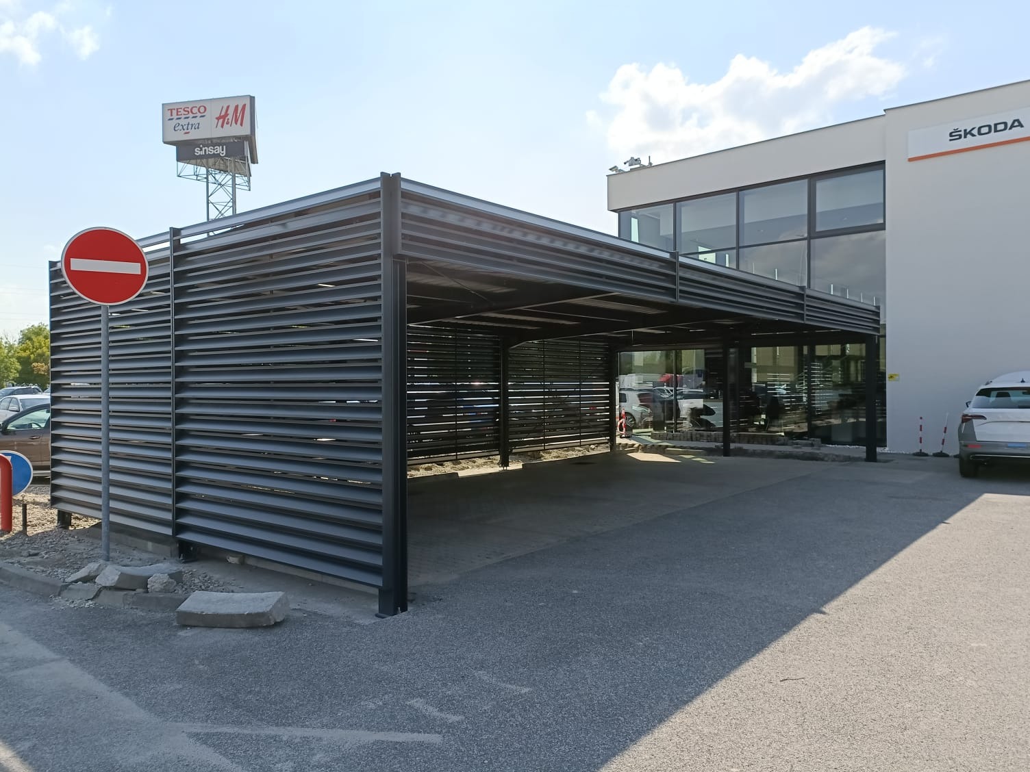Zadaszona konstrukcja stalowa z poziomych profili, przylegająca do budynku salonu samochodowego Skoda, z znakiem zakazu wjazdu po lewej stronie i reklamą Tesco Extra na górze.