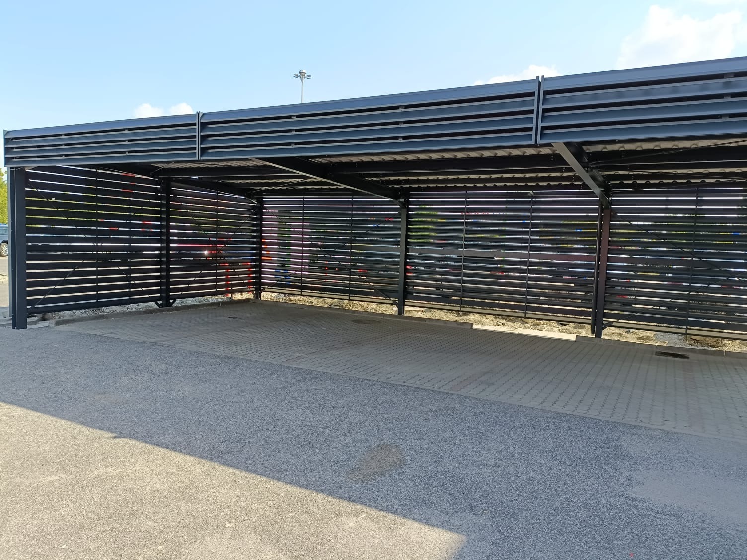 Zadaszenie parkingowe z metalowymi lamelami w kolorze antracytowym, widok z zewnątrz na pusty plac wyłożony kostką brukową, błękitne niebo w tle.