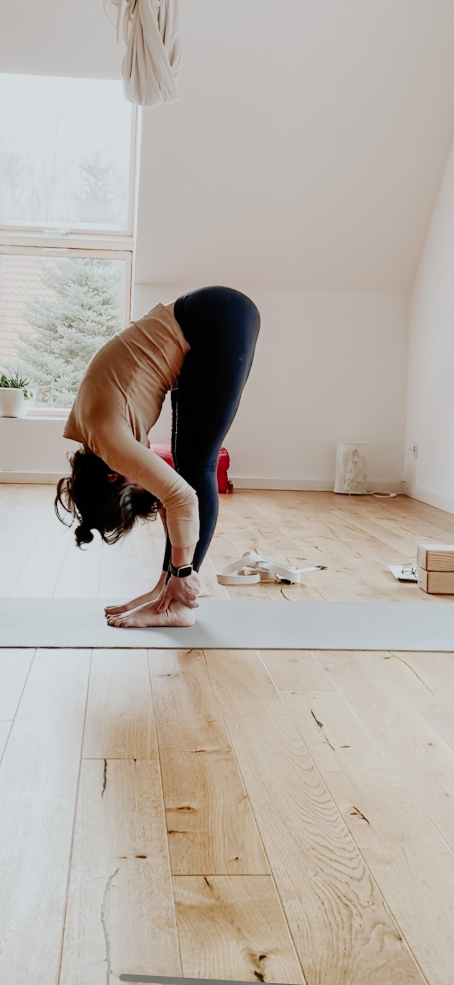 Kobieta w pozycji stojącej, głęboki skłon do przodu, dotyka dłońmi stóp na macie w jasnym pomieszczeniu z drewnianą podłogą i oknem.