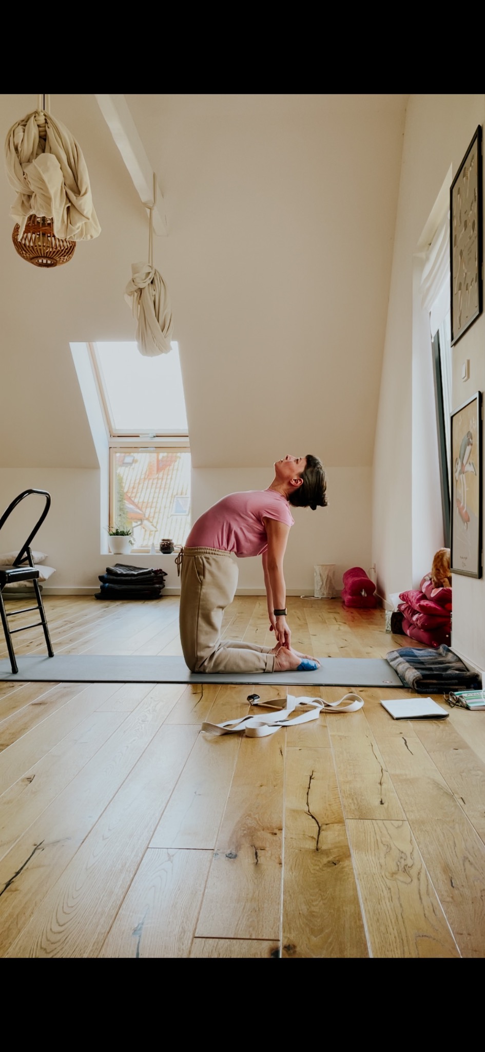 Kobieta w różowej koszulce i beżowych spodniach wykonuje pozycję wielbłąda na macie do ćwiczeń w jasnym pokoju na poddaszu z drewnianą podłogą i oknem dachowym.