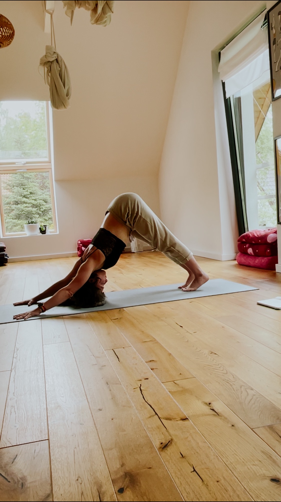 Kobieta w pozycji psa z głową w dół na macie w jasnym pomieszczeniu z drewnianą podłogą i oknami.