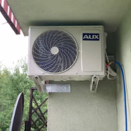 Zewnętrzna jednostka klimatyzatora marki AUX zamontowana na ścianie budynku z widocznymi przewodami i elementami montażowymi, z fragmentem anteny satelitarnej w tle.