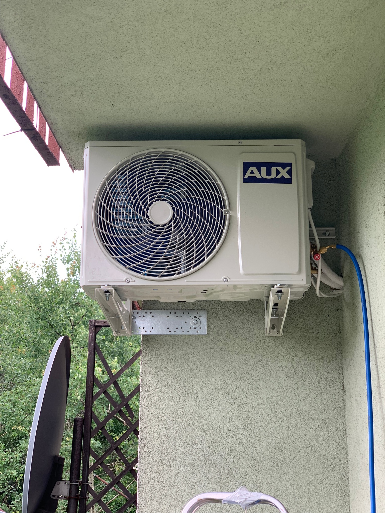 Zewnętrzna jednostka klimatyzatora marki AUX zamontowana na ścianie budynku z widocznymi przewodami i elementami montażowymi, z fragmentem anteny satelitarnej w tle.