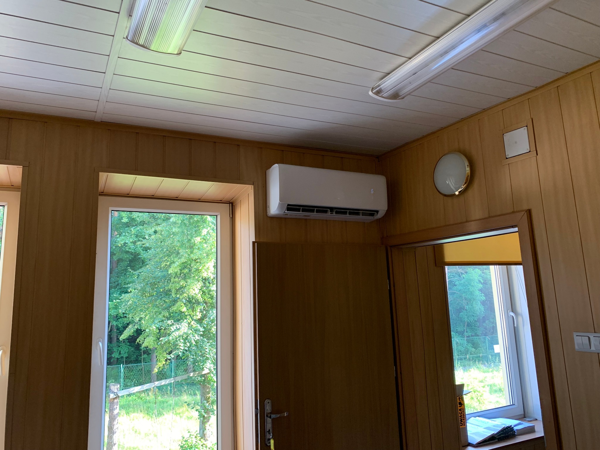 Biały klimatyzator ścienny zamontowany w pomieszczeniu z boazerią, widok z dołu, okno z widokiem na zieleń na zewnątrz.