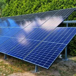 Panel słoneczny z metalową konstrukcją montażową na trawiastym terenie, z odbiciem nieba na powierzchni paneli i żywopłotem w tle.