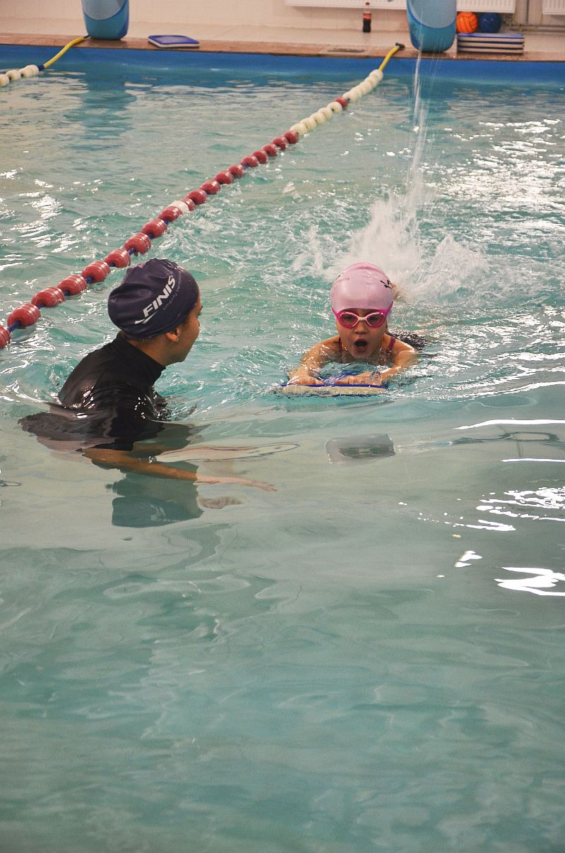 Instruktor w czepku marki Finis obserwuje dziewczynkę w różowym czepku i okularach, która pływa w basenie z deską, z widocznymi pluskami wody.