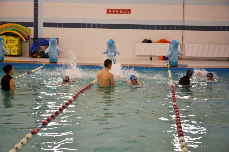 Instruktor pływania obserwuje grupę dzieci podczas lekcji na basenie, z dmuchanymi delfinami dekorującymi brzeg i tabliczką 'Nie biegaj' w tle.